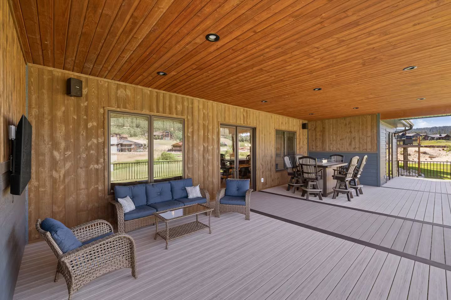 Long covered deck with wicker lounge seating, an outdoor TV and a bar-height fire table with six swivel chairs