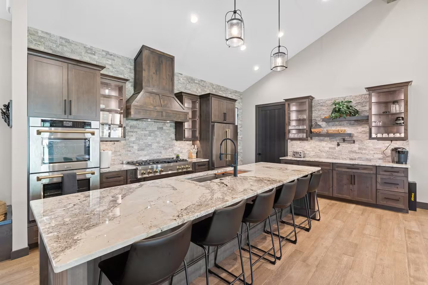 Kitchen with a long stone island and six barstools, double wall ovens, a gas rangetop with brass knobs, a wood hood and a stacked-stone wall