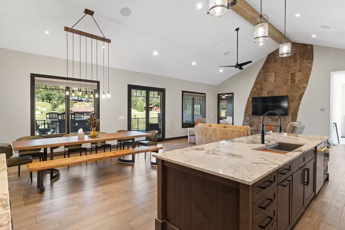 Open main floor at the Luxury Golf Course Retreat: a live-edge dining table with a matching bench, the kitchen island, and the great room with a curved, bark-textured feature wall under a vaulted ceiling