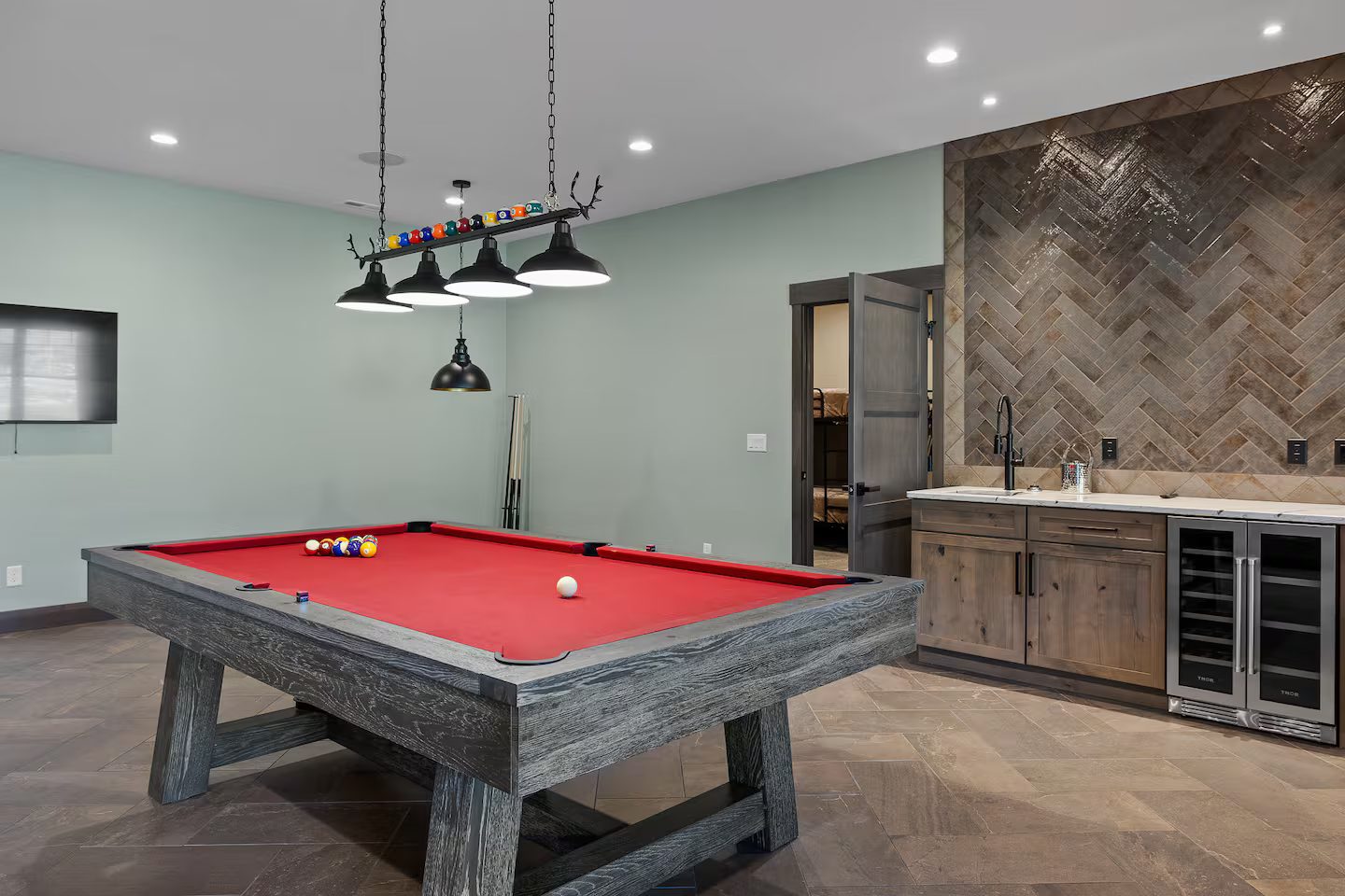 Lower-level game room with a red-felt pool table, billiard lights, a wall TV and a wet bar with a wine fridge